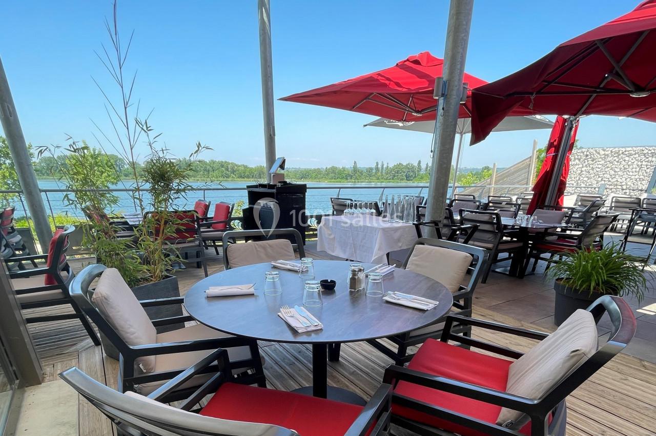 Terrasse d'un restaurant avec tables dressées, chaises rouges et blanches, vue sur un lac et parasols rouges.