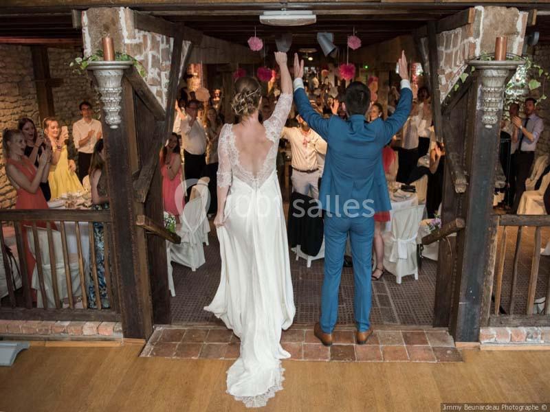 Un couple en tenue de mariage salue des invités dans une salle de réception rustique avec des murs en pierre.