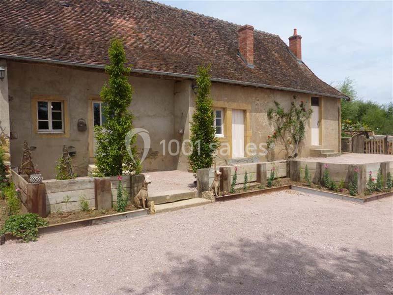 Façade d'une maison ancienne en pierre avec toit en tuiles, jardinières en bois et allée gravillonnée au premier plan.