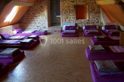 Dortoir sous combles avec murs en pierre, matelas au sol alignés et poutres apparentes.