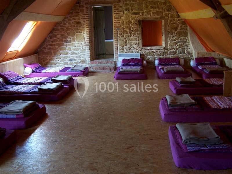 Dortoir mansardé avec matelas violets alignés au sol, murs en pierre apparente et lumière naturelle.