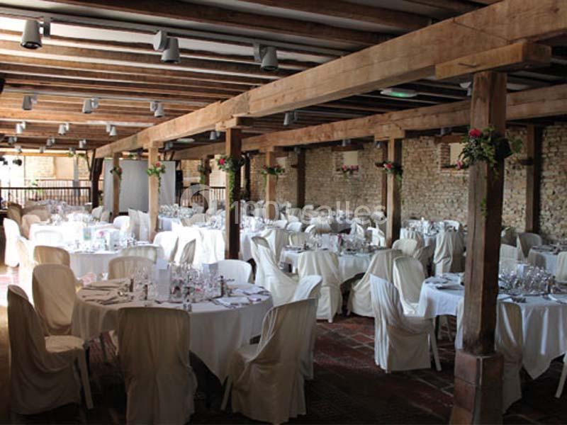 Salle de réception avec des tables rondes dressées, nappes blanches et poutres apparentes en bois.