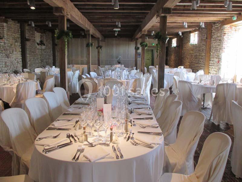 Salle de réception avec tables rondes et rectangulaires dressées, chaises couvertes de housses blanches et poutres…