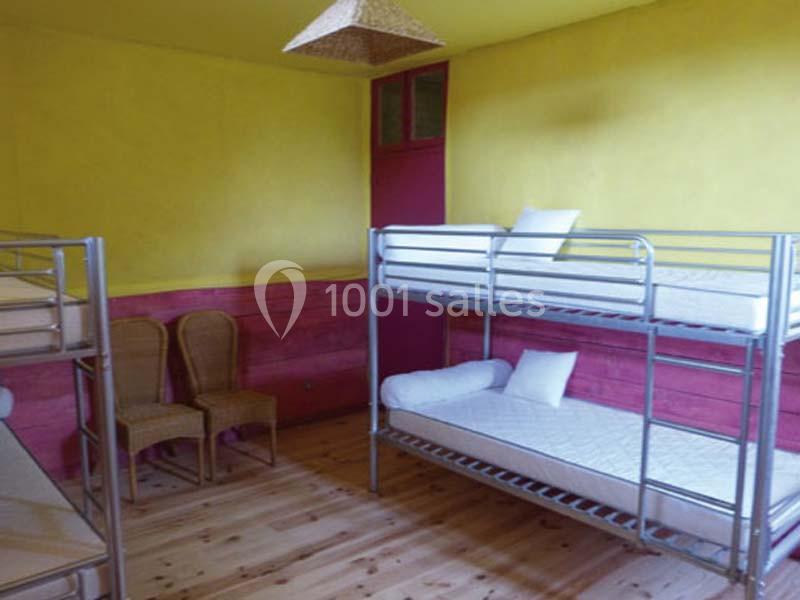 Chambre avec lits superposés en métal, murs jaunes et roses, sol en bois, deux chaises en osier et un plafonnier.