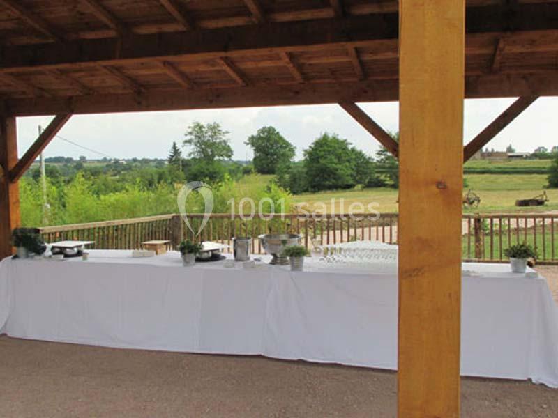 Table dressée sous un abri en bois, avec nappes blanches, vaisselle et verdure, donnant sur un paysage champêtre.