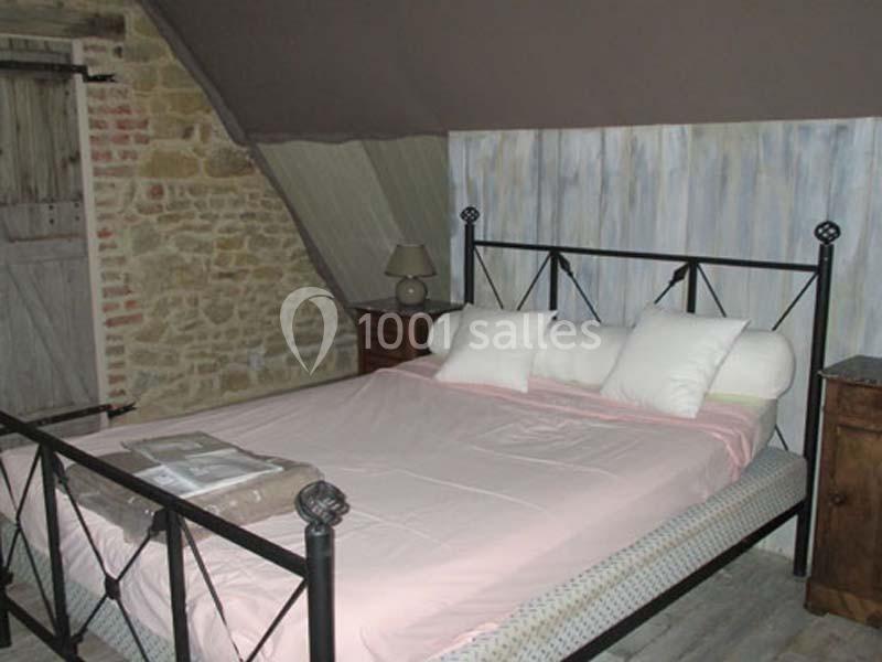 Lit double avec cadre en métal noir, draps roses, oreillers blancs, dans une chambre aux murs en pierre et bois.