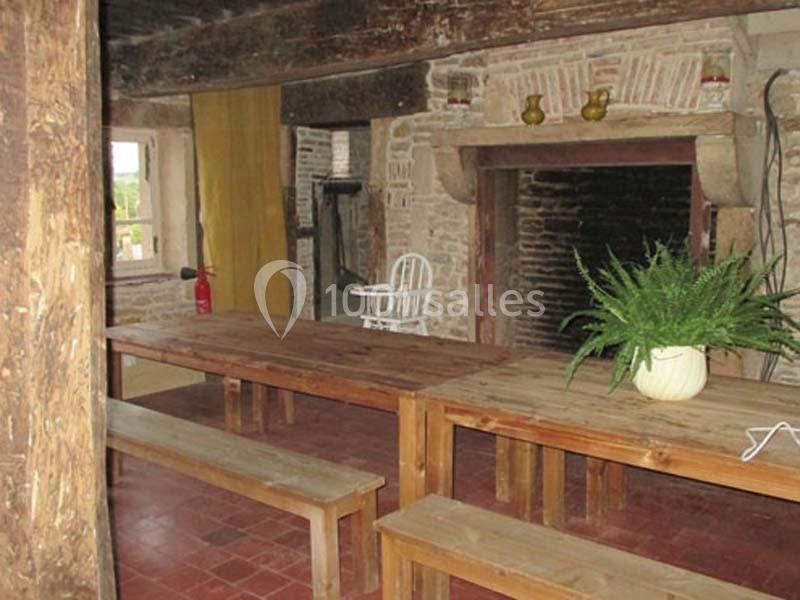 Salle rustique avec murs en pierre, poutres apparentes, grande cheminée, tables en bois et une plante en pot.