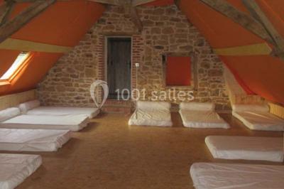 Dortoir sous combles avec murs en pierre, matelas au sol alignés et poutres apparentes.