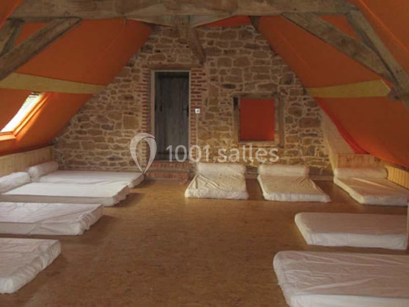 Dortoir sous combles avec murs en pierre, matelas au sol alignés et poutres apparentes.