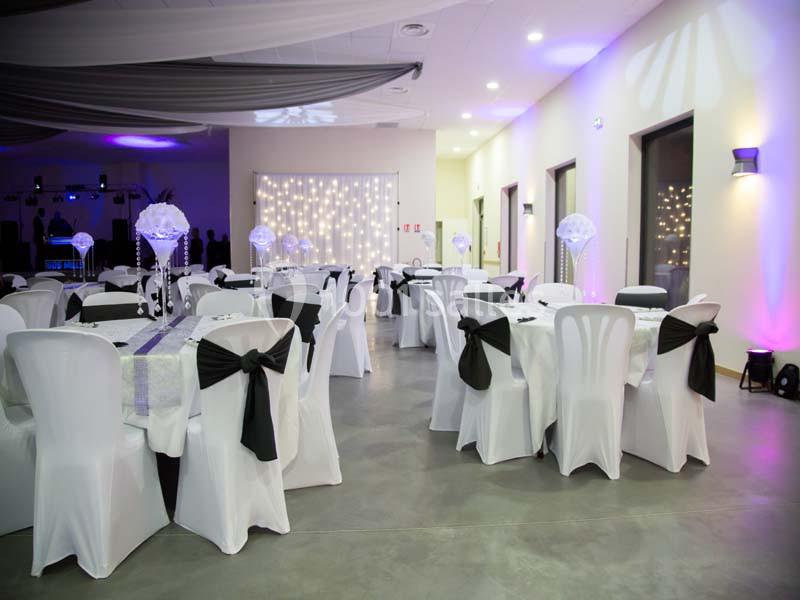 Salle de réception décorée avec des tables rondes, nappes blanches, chaises couvertes et éclairage tamisé.