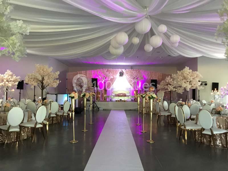 Salle de réception décorée avec des tables rondes, des chaises blanches, des arbres artificiels et un éclairage rose.