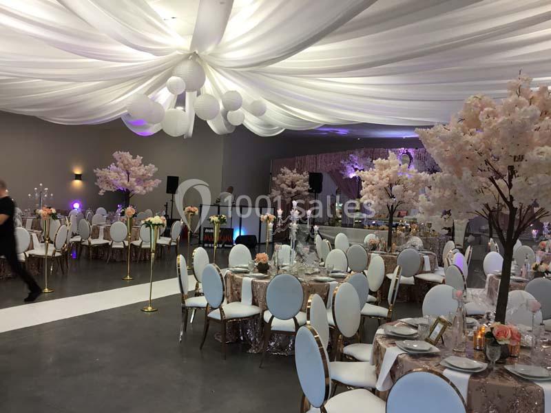 Salle de réception décorée avec des tables élégantes, des chaises blanches, des arbres fleuris et un plafond drapé.