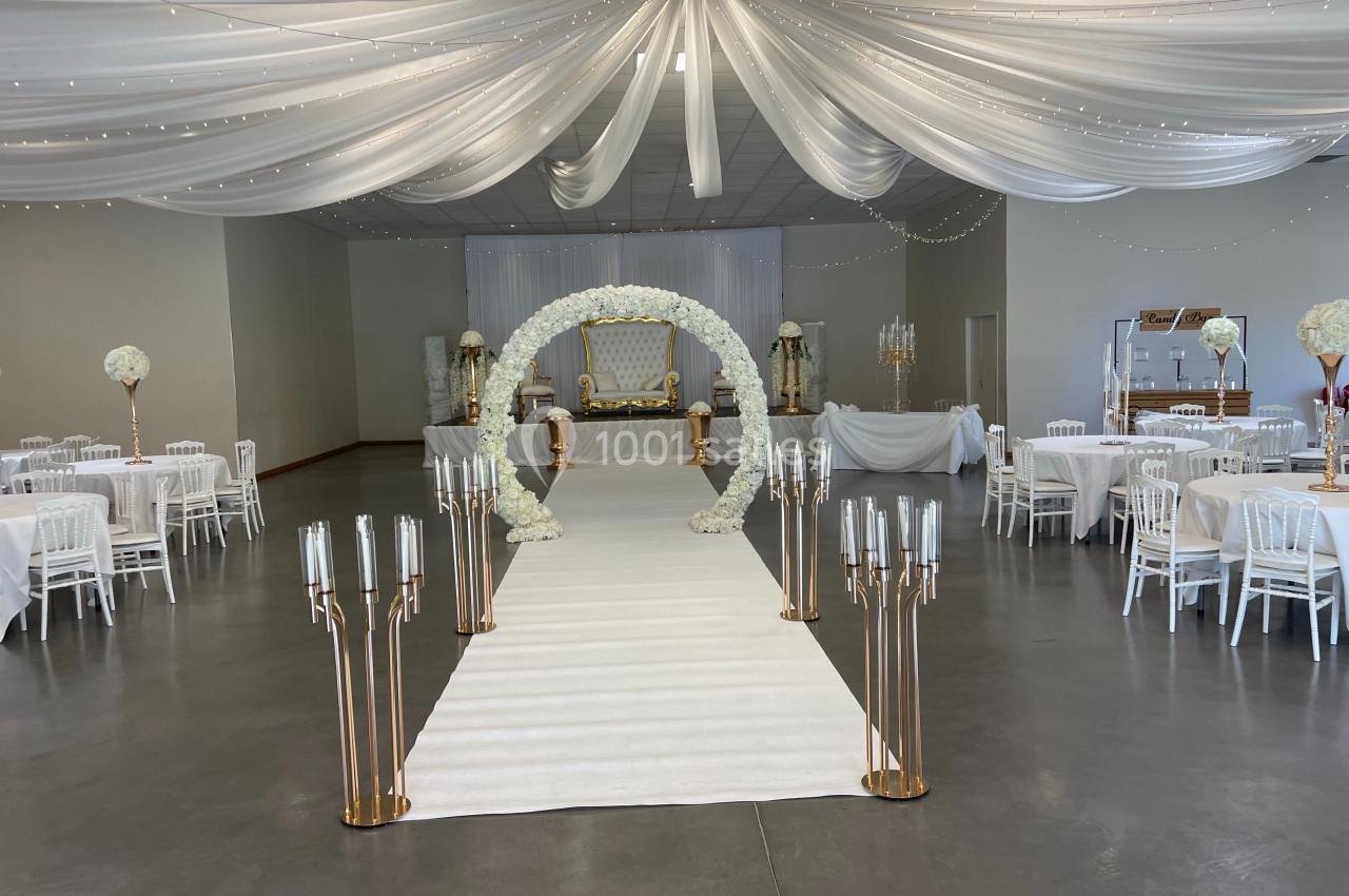 Salle de réception décorée avec des drapés blancs, une arche florale, un tapis blanc et des chandeliers dorés.