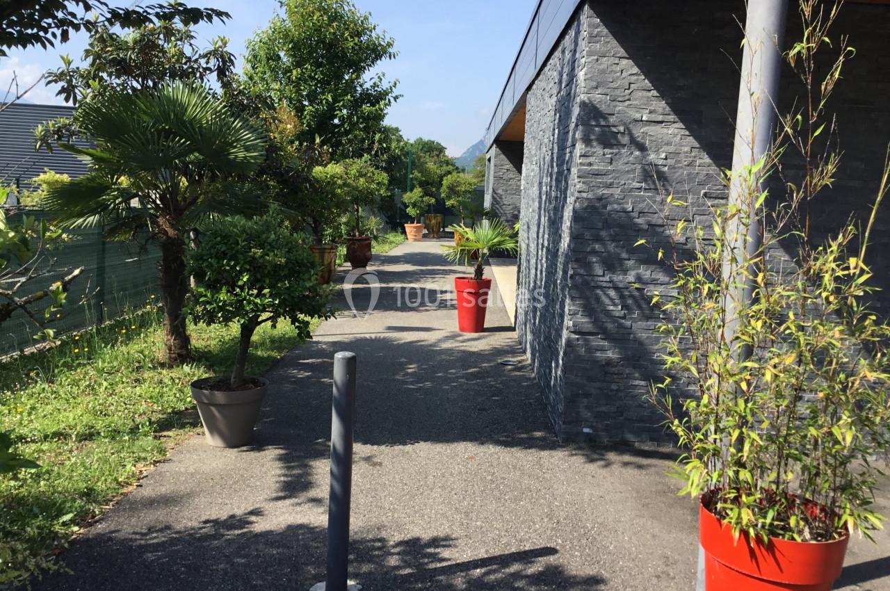 Allée extérieure bordée de plantes en pots, longeant un bâtiment aux murs en pierre grise sous un ciel dégagé.