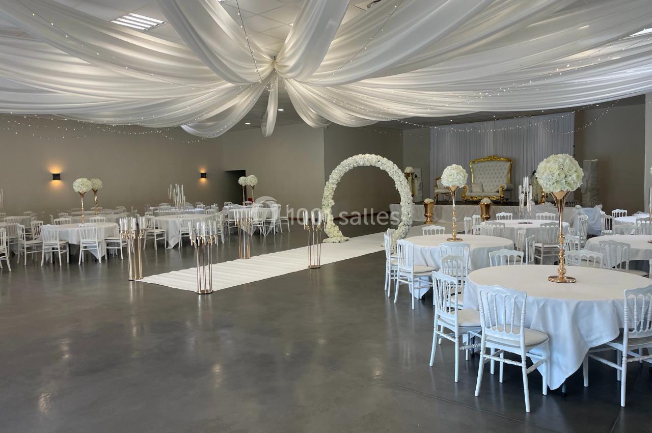 Salle de réception décorée avec des tables rondes, des chaises blanches, des fleurs et un plafond drapé de tissu blanc.