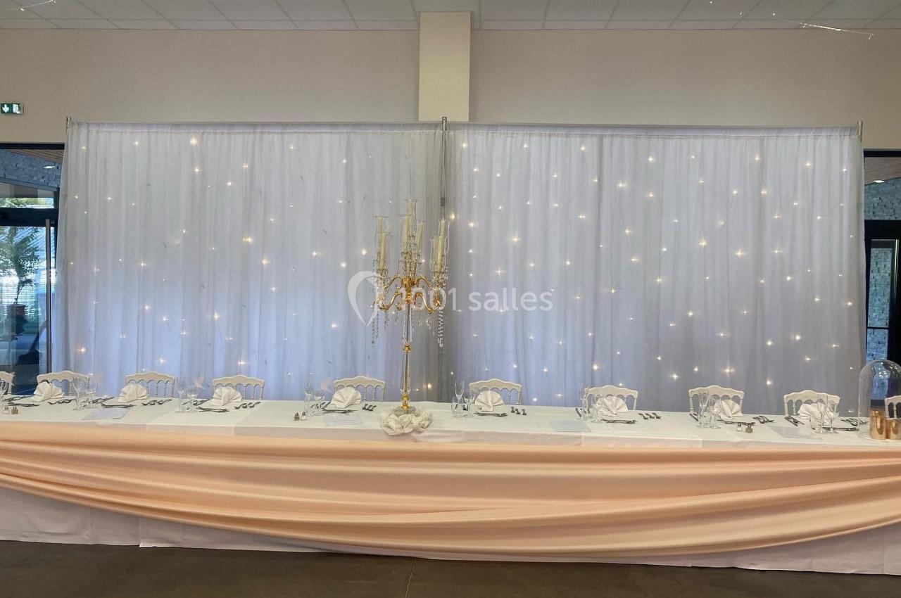 Table de banquet décorée avec nappes blanches, chandeliers dorés et fond en rideaux lumineux.