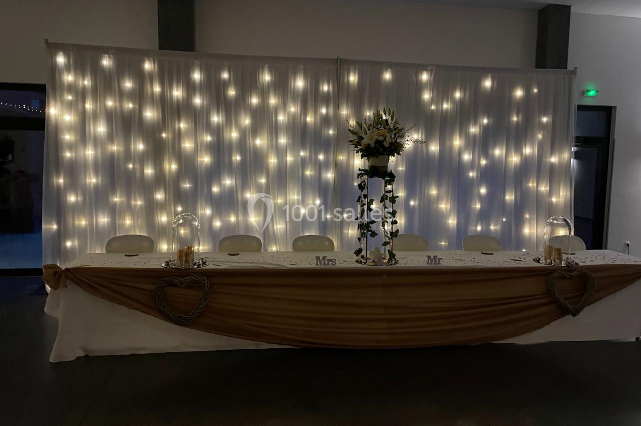 Table de mariage décorée avec une nappe dorée, des fleurs, des bougies et un fond lumineux de guirlandes LED.