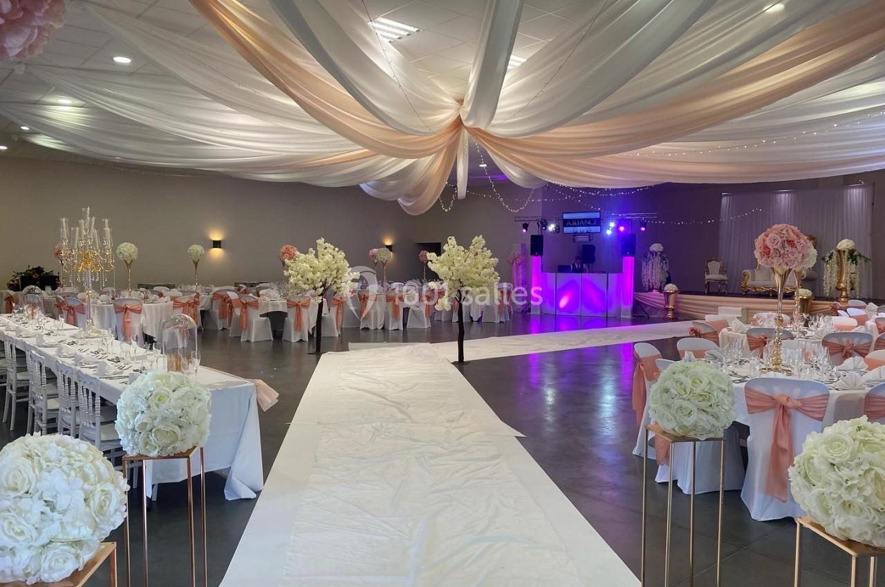 Salle de réception décorée avec des drapés blancs, tables dressées, fleurs blanches et roses, et éclairage tamisé.