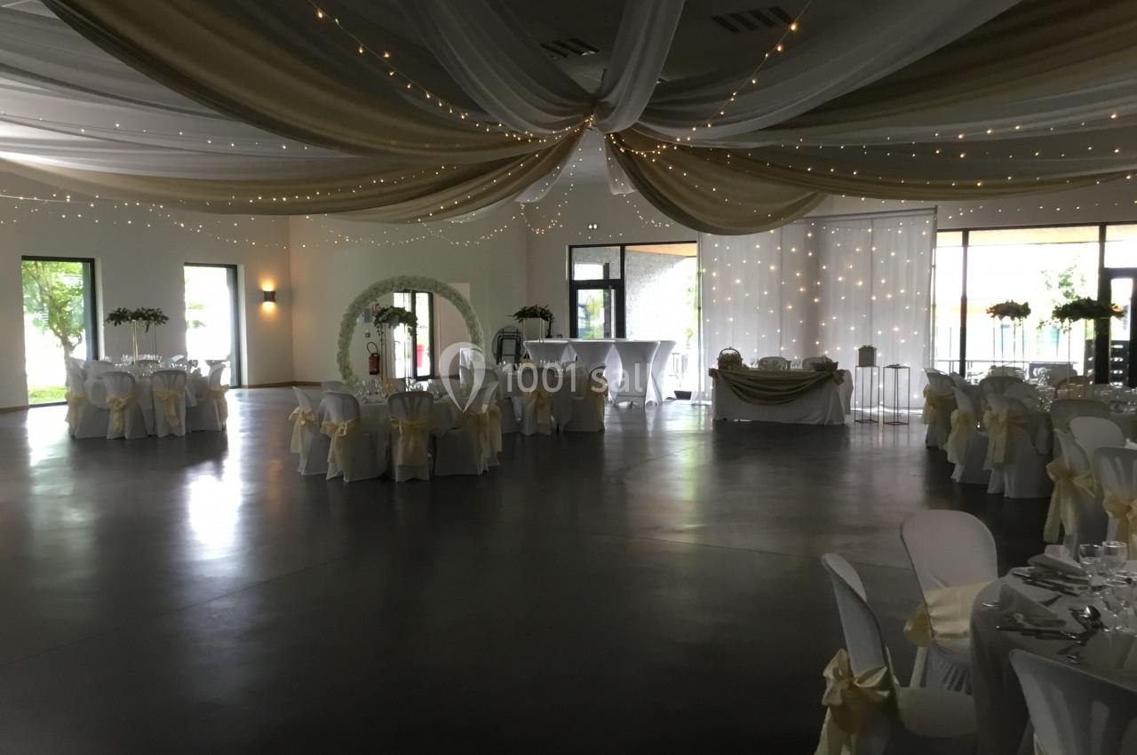 Salle de réception décorée avec des tables rondes, des chaises ornées de nœuds et un plafond drapé de tissus et guirlandes…