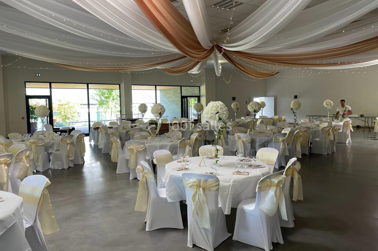 Salle de réception décorée avec des tables rondes, nappes blanches, chaises ornées de rubans dorés et plafonds drapés.