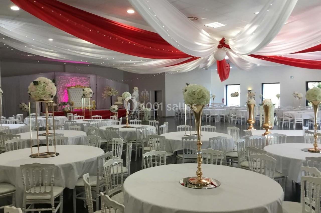 Salle de réception décorée avec des tables rondes, des chaises blanches et des drapés rouges et blancs au plafond.