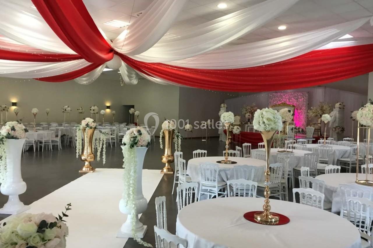 Salle de réception décorée avec des drapés rouges et blancs au plafond, tables rondes et arrangements floraux élégants.