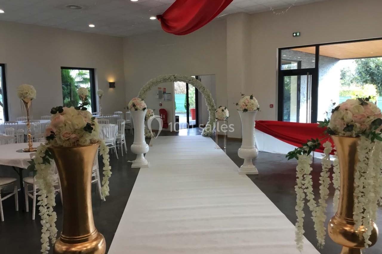 Salle de réception décorée pour un mariage avec arche florale, tapis blanc et vases dorés ornés de fleurs.