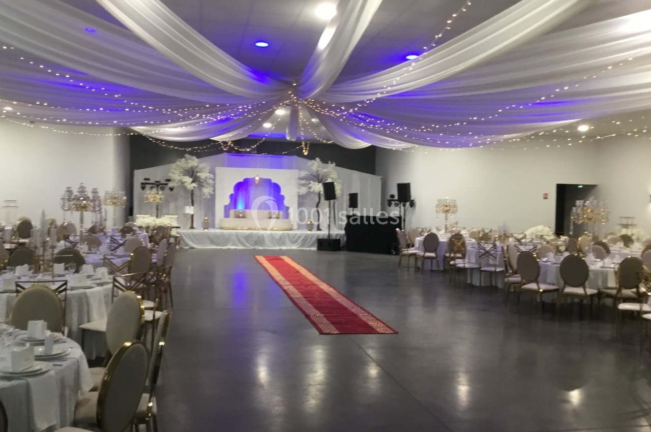 Salle de réception décorée avec des tables rondes, des chaises dorées, un tapis rouge et un éclairage blanc et violet.