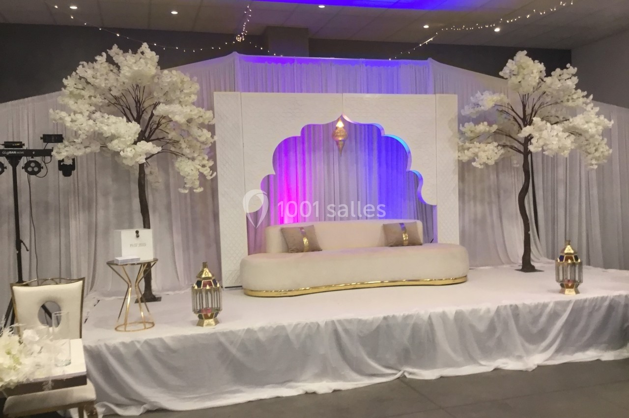 Décor de scène avec canapé beige, lanternes dorées, rideaux blancs et arbres artificiels ornés de fleurs blanches.