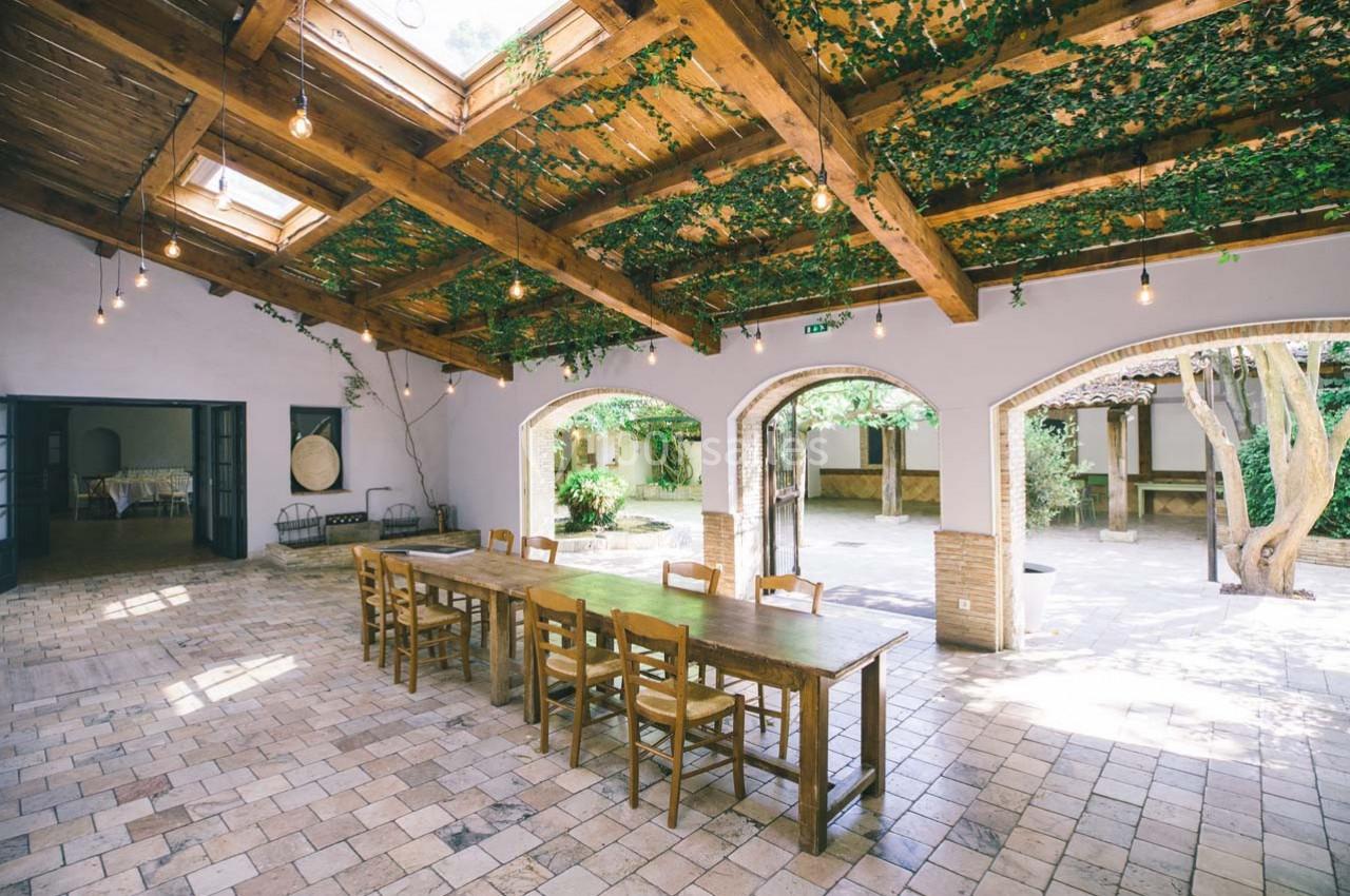 Salle lumineuse avec toit en bois, plantes suspendues, grande table en bois et vue sur une cour arborée.
