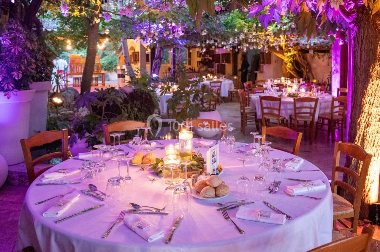 Table ronde dressée pour un dîner en extérieur, entourée de chaises, sous des guirlandes lumineuses et végétation.