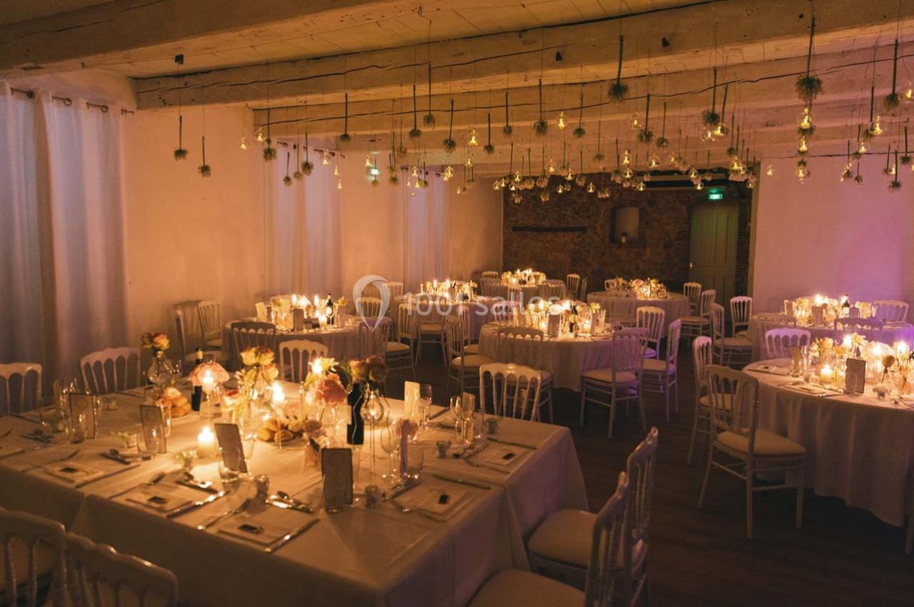 Salle de réception décorée avec des tables rondes, nappes blanches, bougies et suspensions lumineuses.