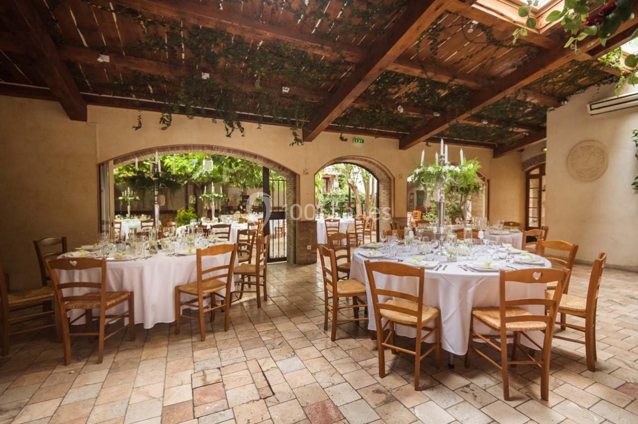 Salle de réception lumineuse avec tables rondes dressées, chaises en bois et plafond décoré de végétation suspendue.