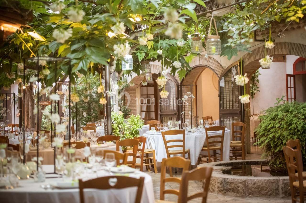 Terrasse d'un restaurant avec des tables dressées, entourée de verdure et éclairée par des lanternes suspendues.