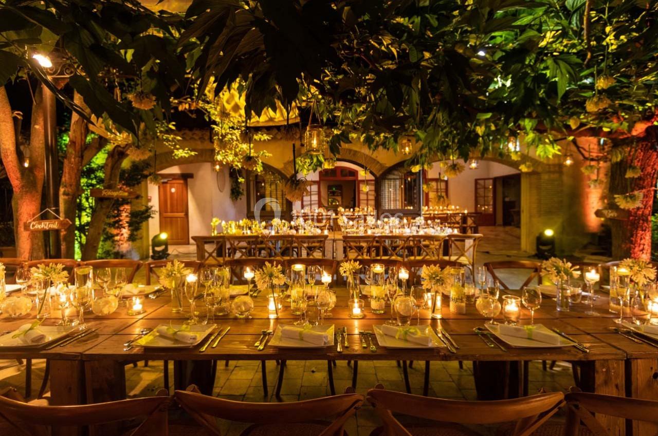 Tables en bois décorées de bougies et de fleurs jaunes, disposées dans un jardin éclairé pour un dîner en soirée.