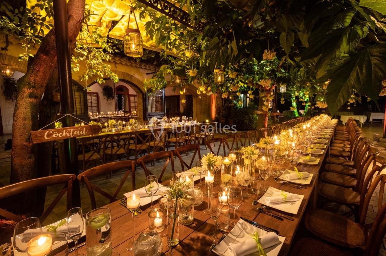 Grande table en bois décorée de bougies et fleurs, installée dans un jardin éclairé pour un dîner en soirée.