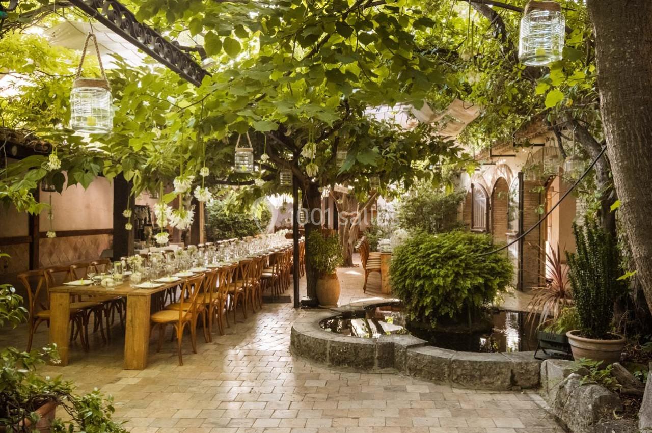 Salle à manger extérieure sous une pergola végétalisée, avec une longue table en bois et des plantes luxuriantes autour.