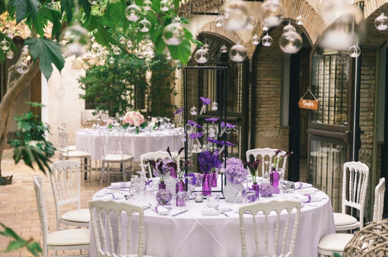 Table décorée avec des tons violets dans une cour intérieure, entourée de plantes et de guirlandes lumineuses suspendues.