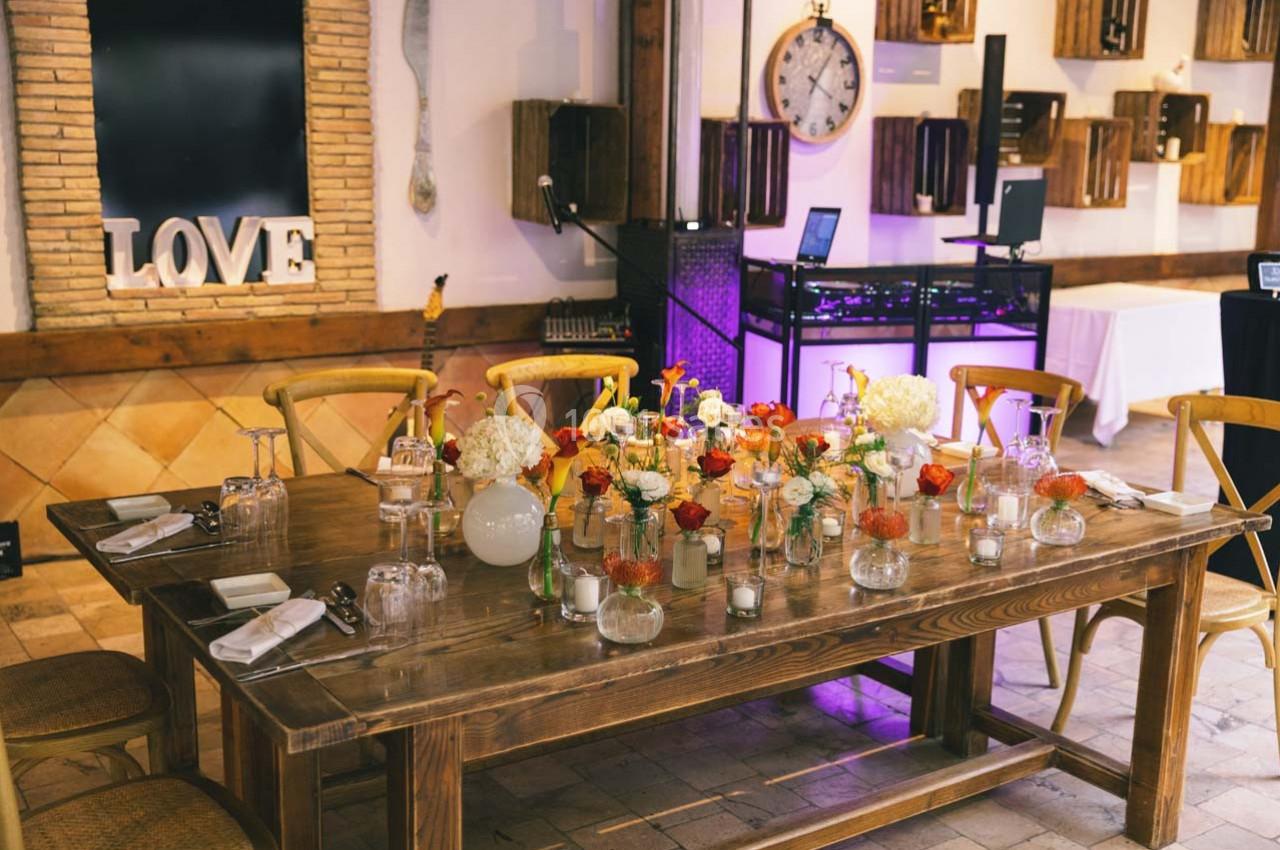Table en bois décorée de fleurs, bougies et vaisselle, dans une salle avec murs en briques et éléments décoratifs.