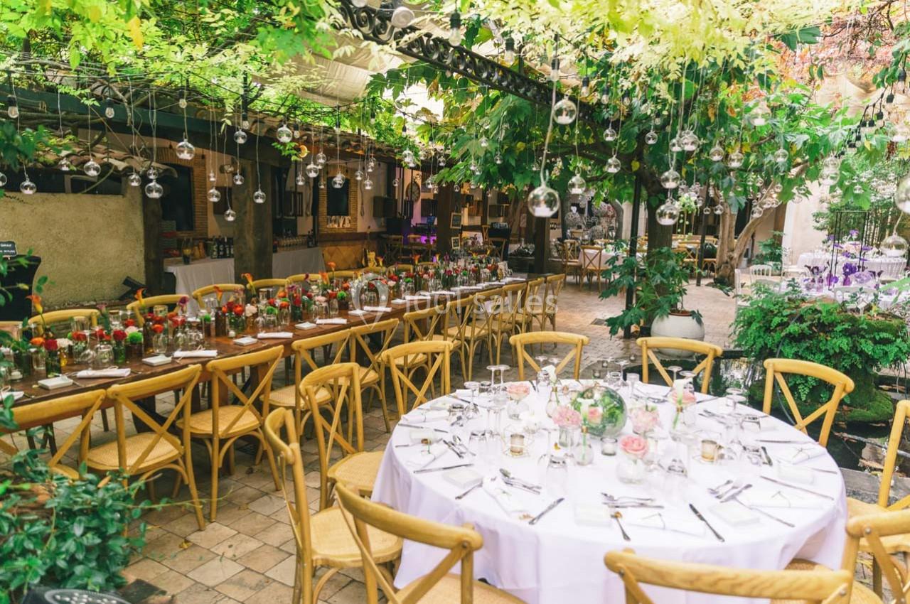 Salle extérieure décorée avec des guirlandes végétales, tables rondes et rectangulaires dressées pour un repas.