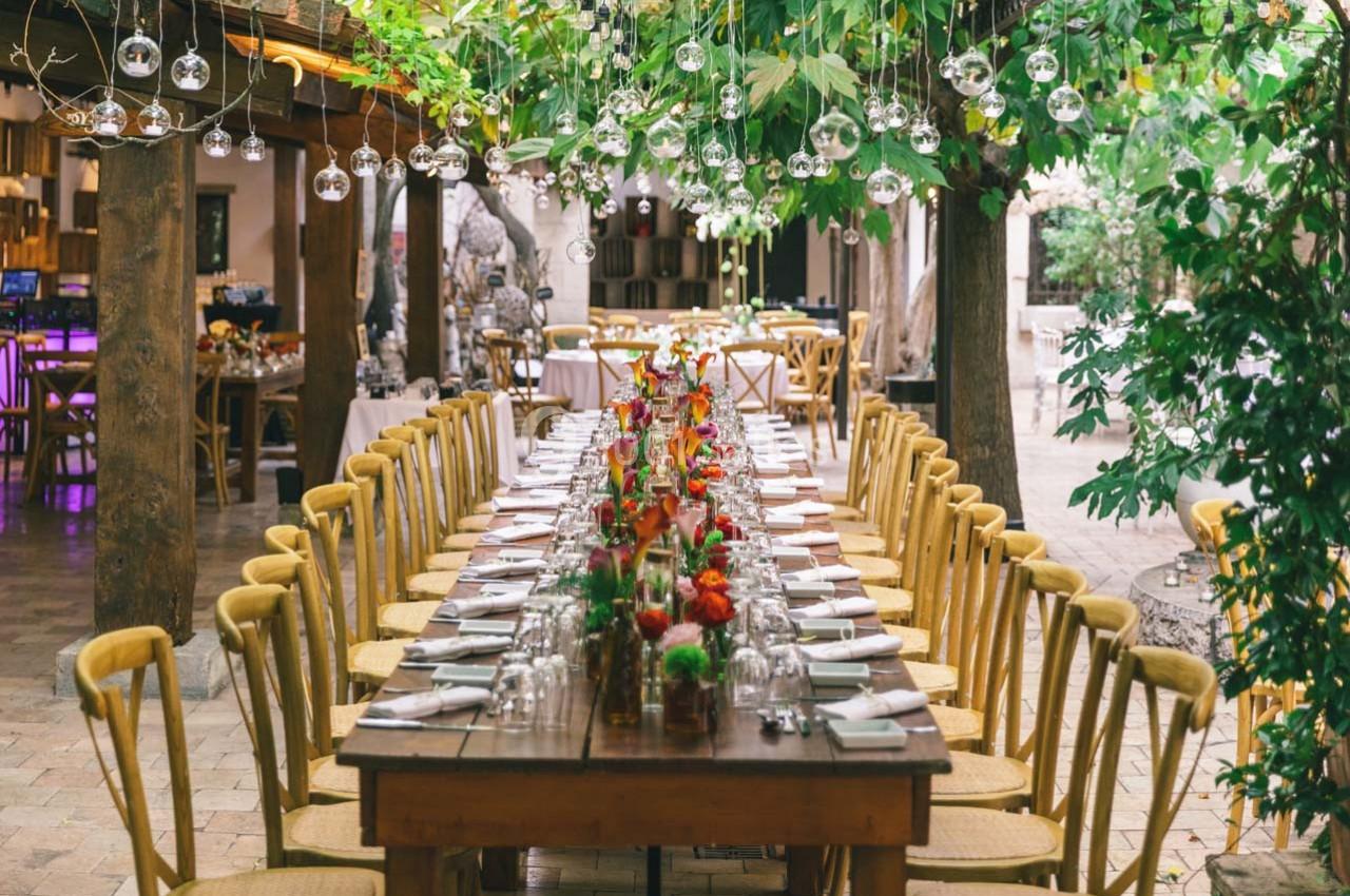 Grande table en bois décorée de fleurs colorées, entourée de chaises, sous un toit végétalisé avec des lumières suspendues.