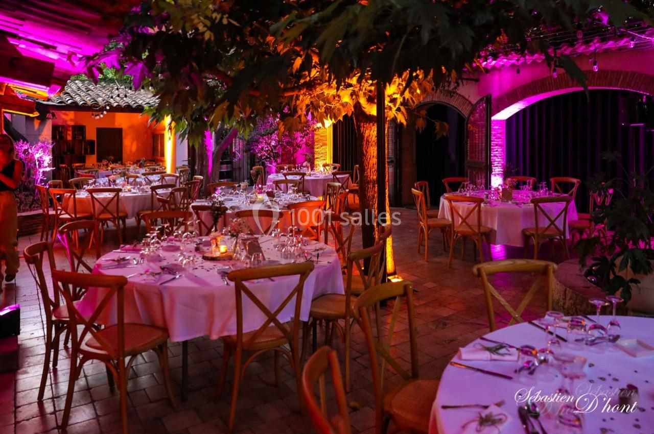 Salle de réception avec tables rondes dressées, éclairage coloré et ambiance chaleureuse sous un toit partiellement ouvert.