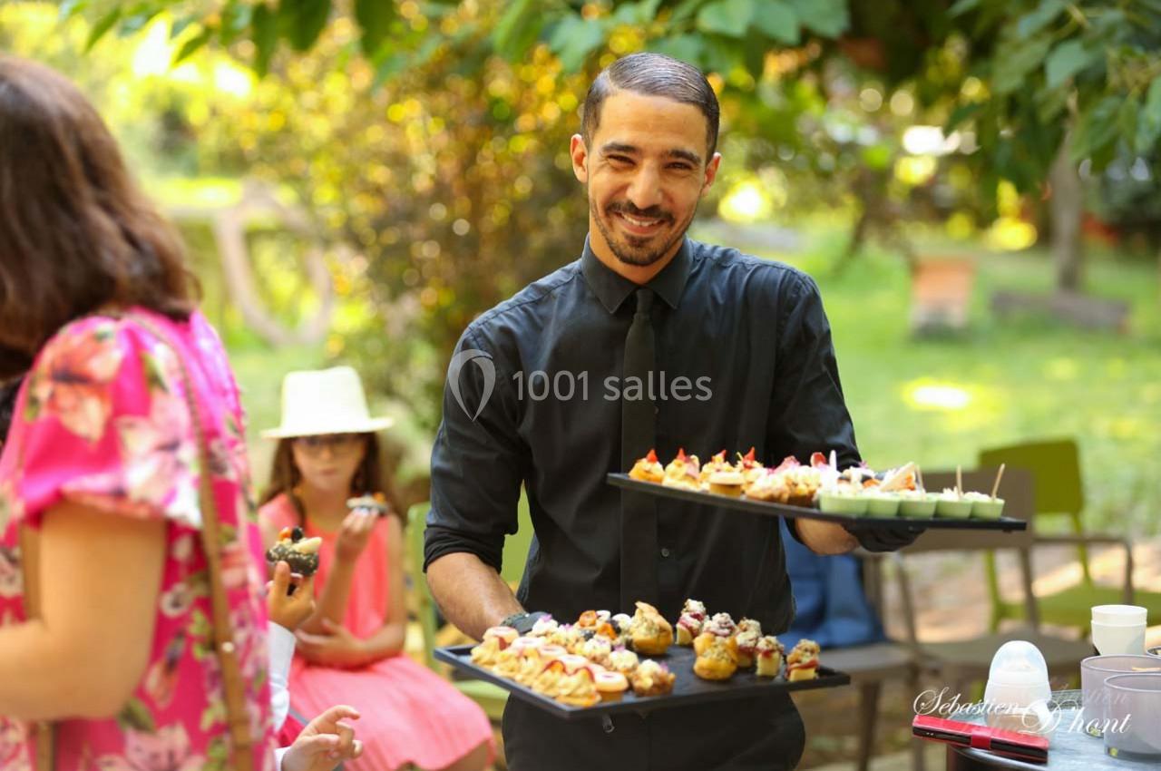 Un serveur souriant présente des plateaux de bouchées apéritives dans un jardin lors d'un événement en plein air.