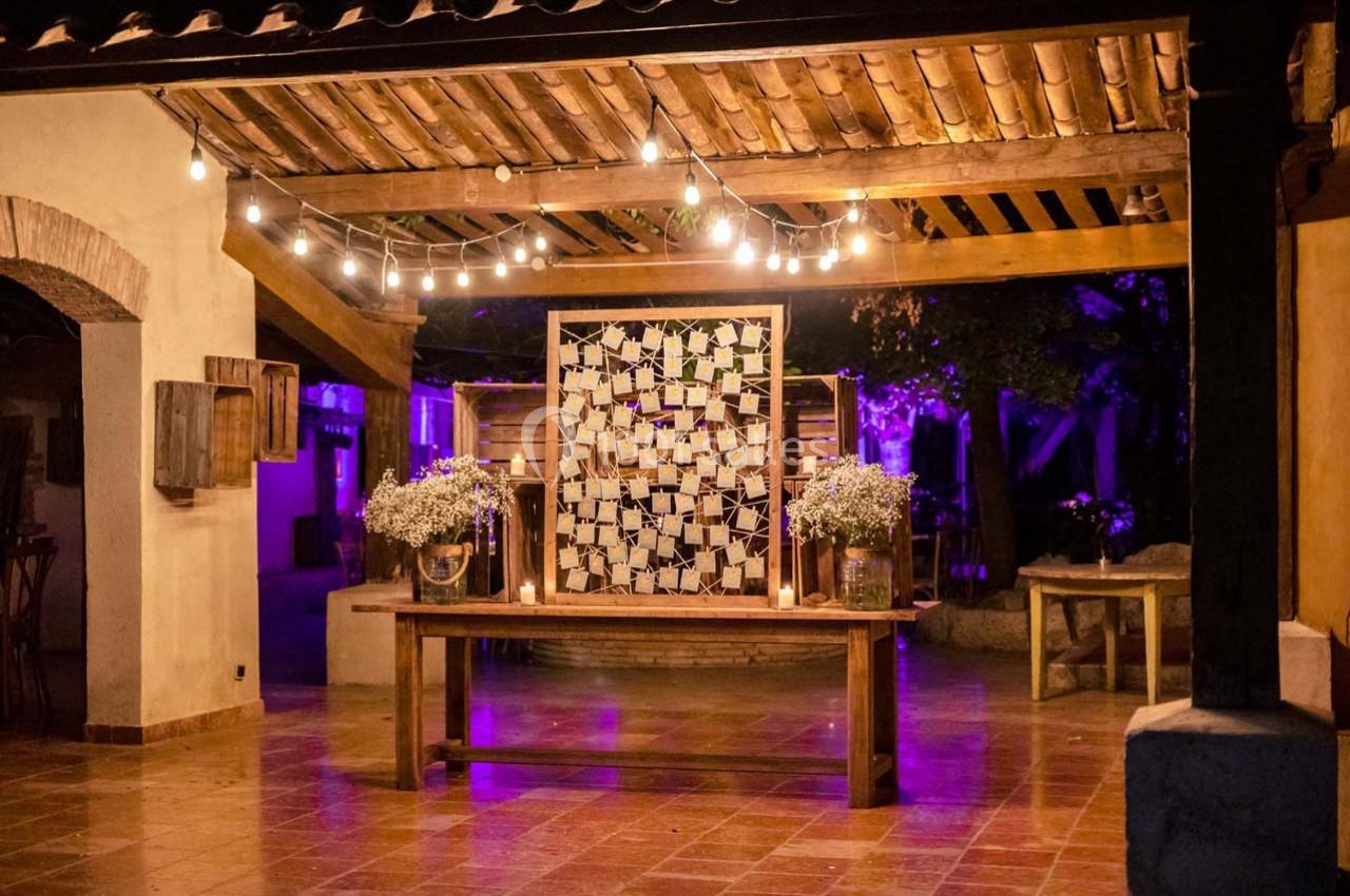 Décoration de mariage en soirée avec guirlandes lumineuses, table en bois et mur orné de photos sous une pergola.