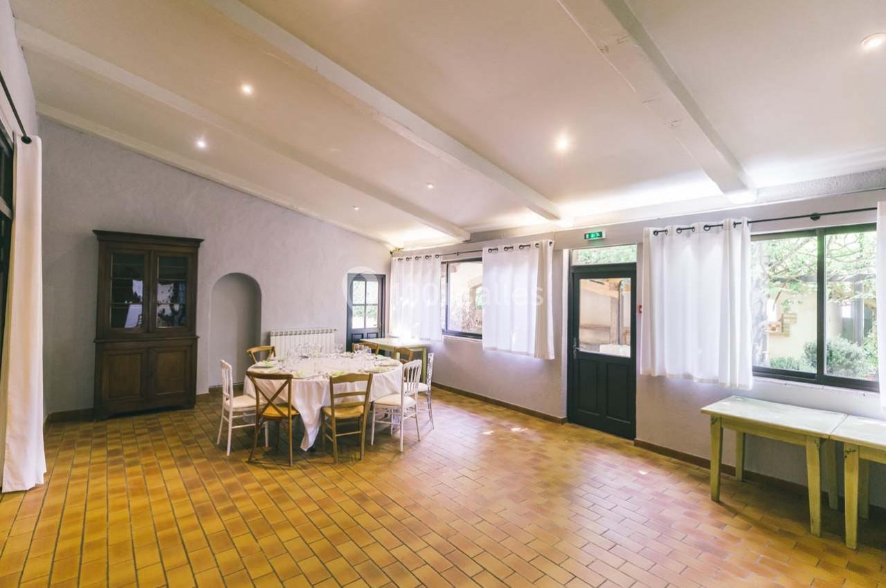 Salle lumineuse avec carrelage en terre cuite, tables dressées, chaises blanches et grandes fenêtres avec rideaux.