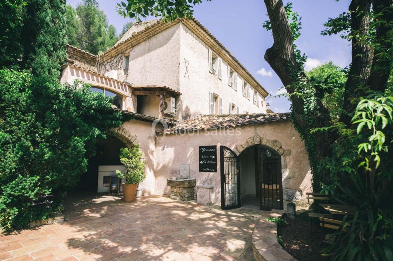 Façade d'un bâtiment en pierre avec toit en tuiles, entouré de végétation et d'une cour pavée ombragée.