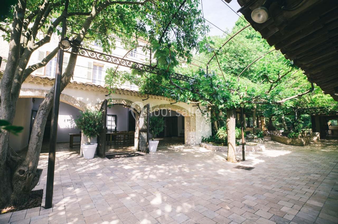 Cour intérieure pavée entourée d'arbres et de végétation, avec une façade en arches et un toit en tuiles.