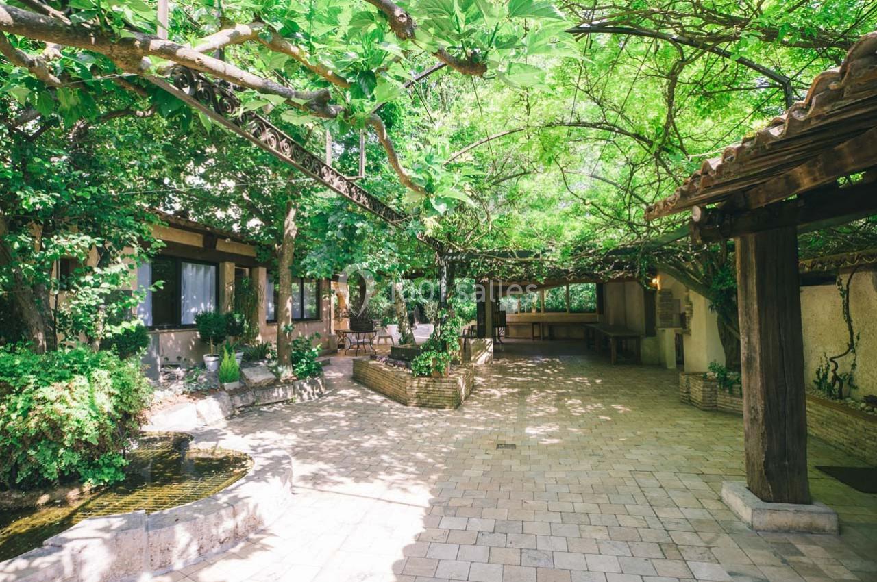 Cour extérieure pavée ombragée par une pergola couverte de végétation, avec un petit bassin et des plantes.