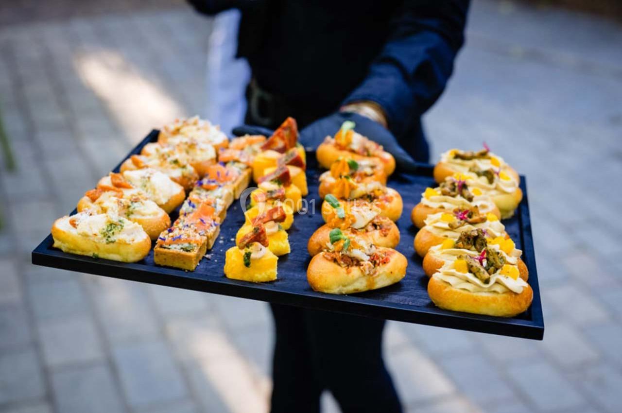 Plateau de canapés variés présentés sur un fond de pavés, tenu par une personne en tenue sombre.