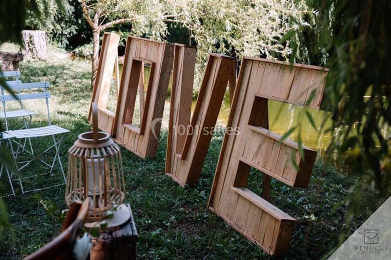 Grandes lettres en bois formant le mot ’LOVE’ disposées sur une pelouse près d'un étang, entourées de verdure.